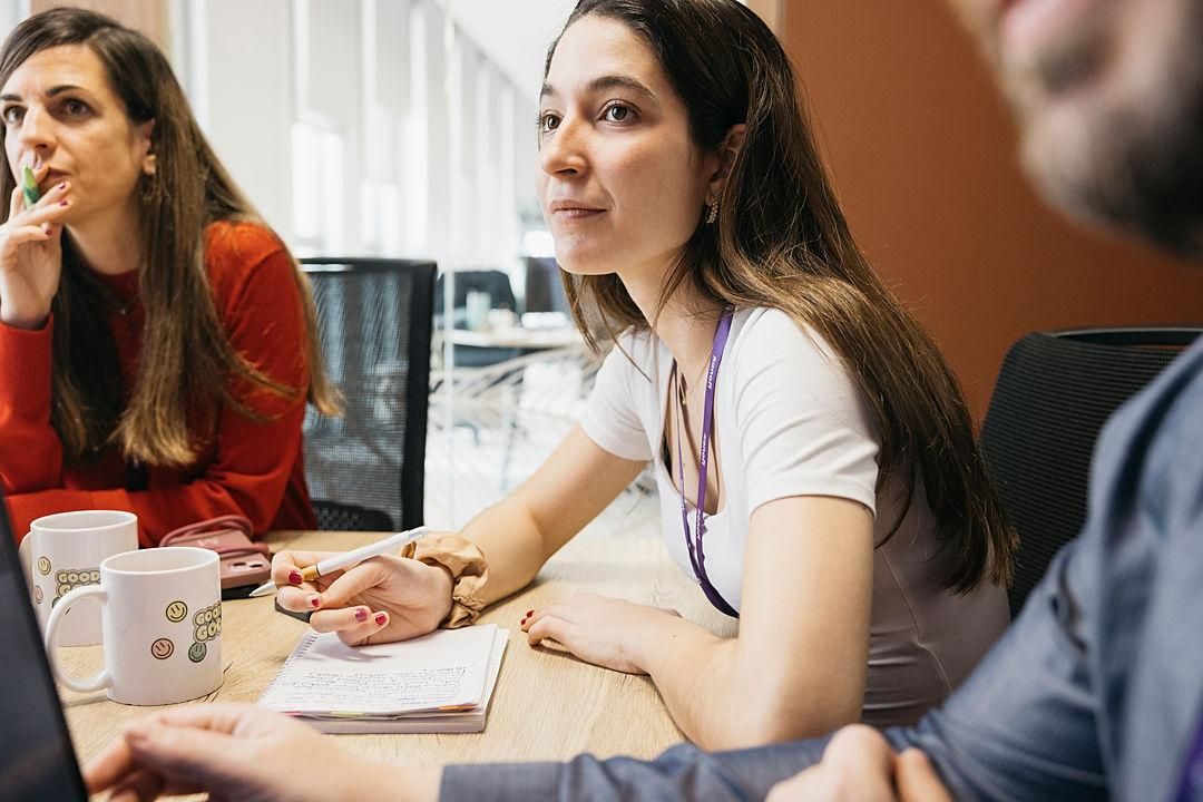 Woman with long hair sitting with colleagues, pen in hand, listening to her interlocutors.