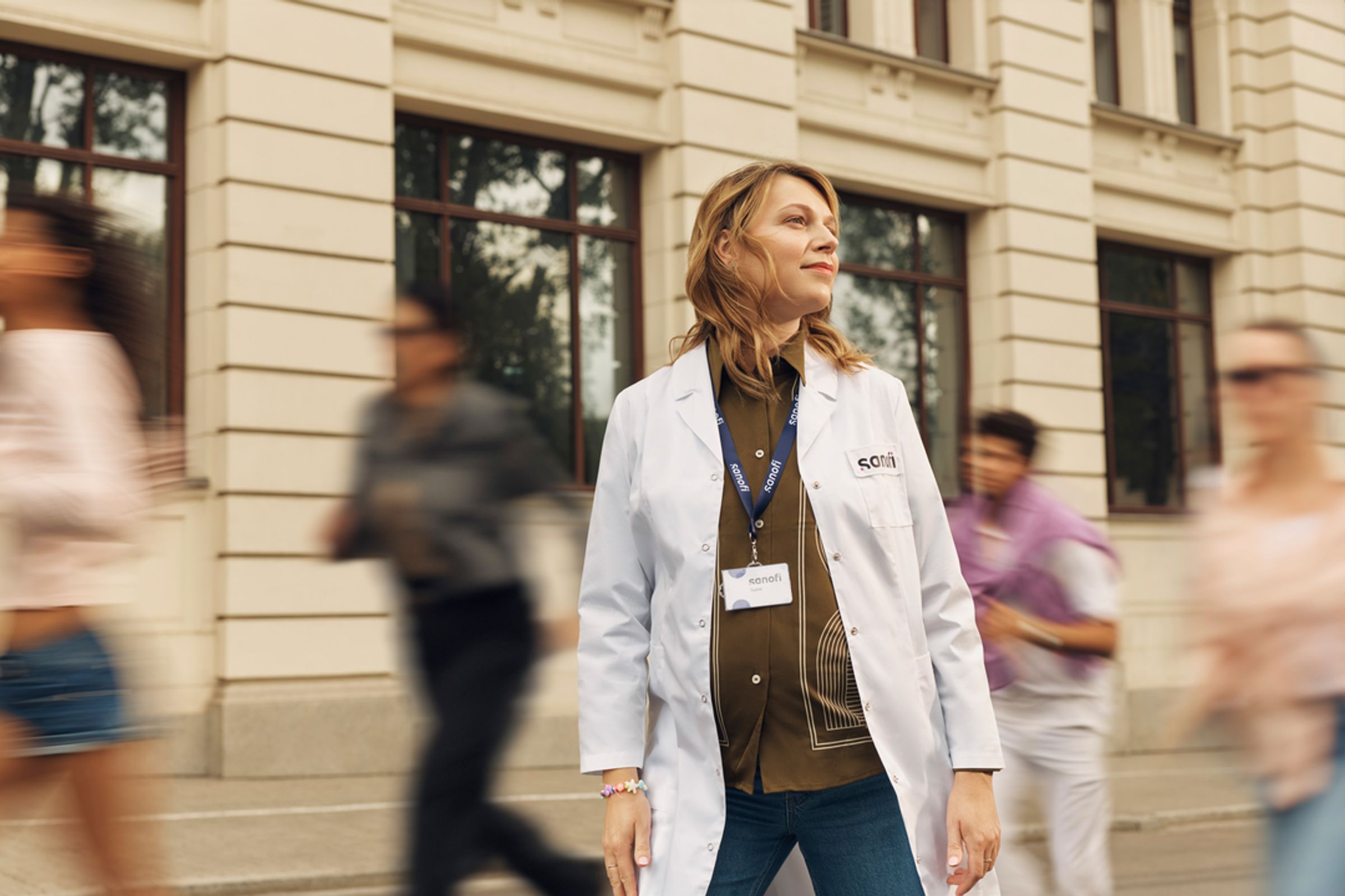 Sanofi employee wearing a white coat and identification badge, walking through an urban environment with pedestrians moving around her.
