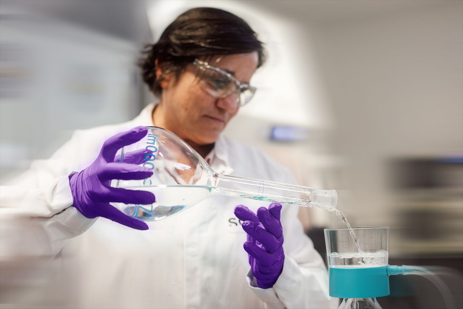 Person wearing a lab coat, safety glasses and gloves, pouring liquid into a filtration device in a laboratory.
