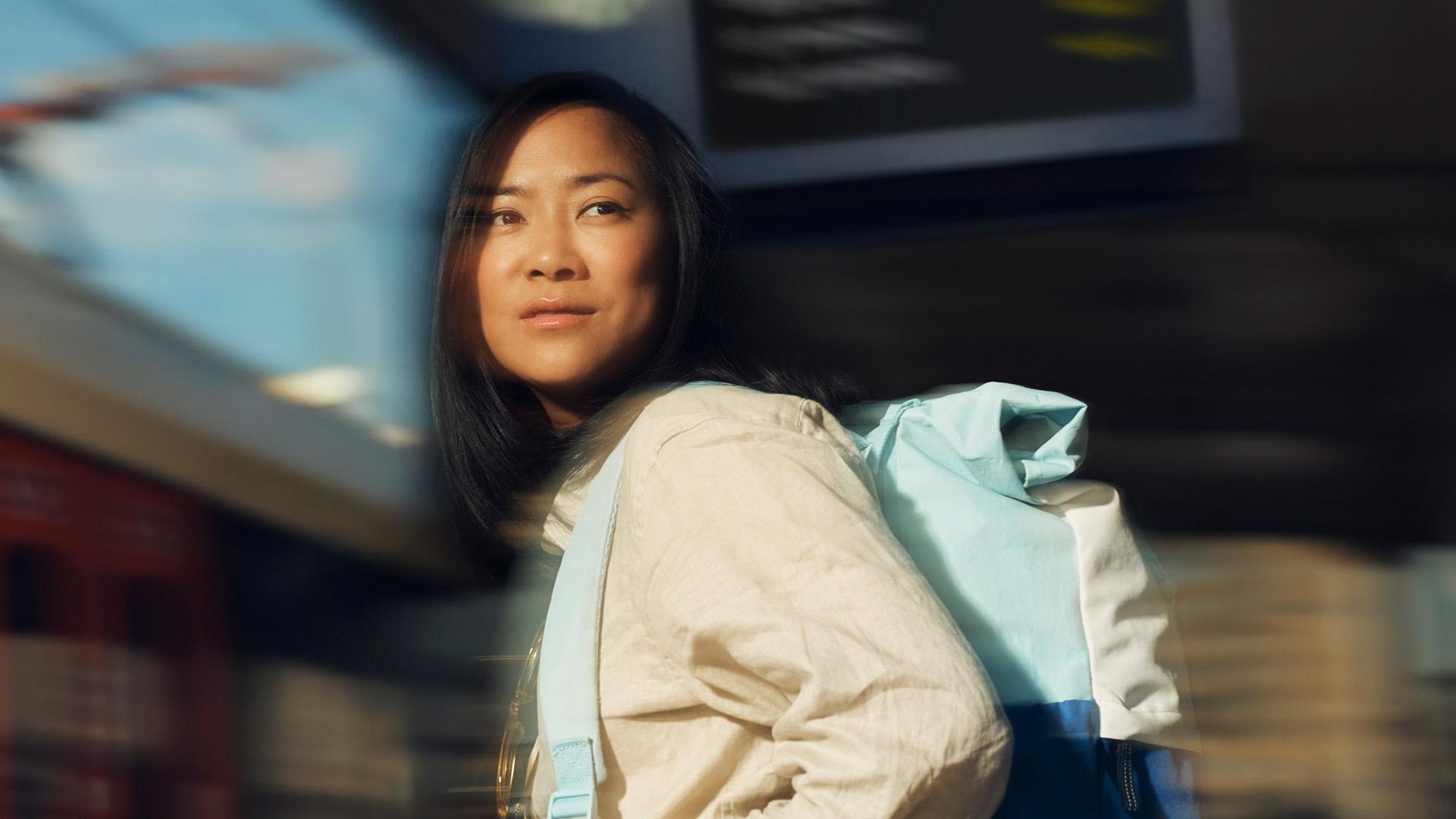 淡い黄色のシャツを着て、青いリュックサックを背負った女性が、駅の中を歩きながら前を見つめている。