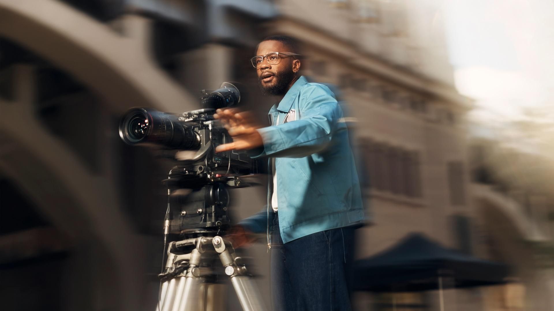 A man wearing glasses and a blue jacket operates a video camera and looks focused while filming, his hand is outstretched in front of him as he gestures. Overlay text reads: "Filmmaker."​