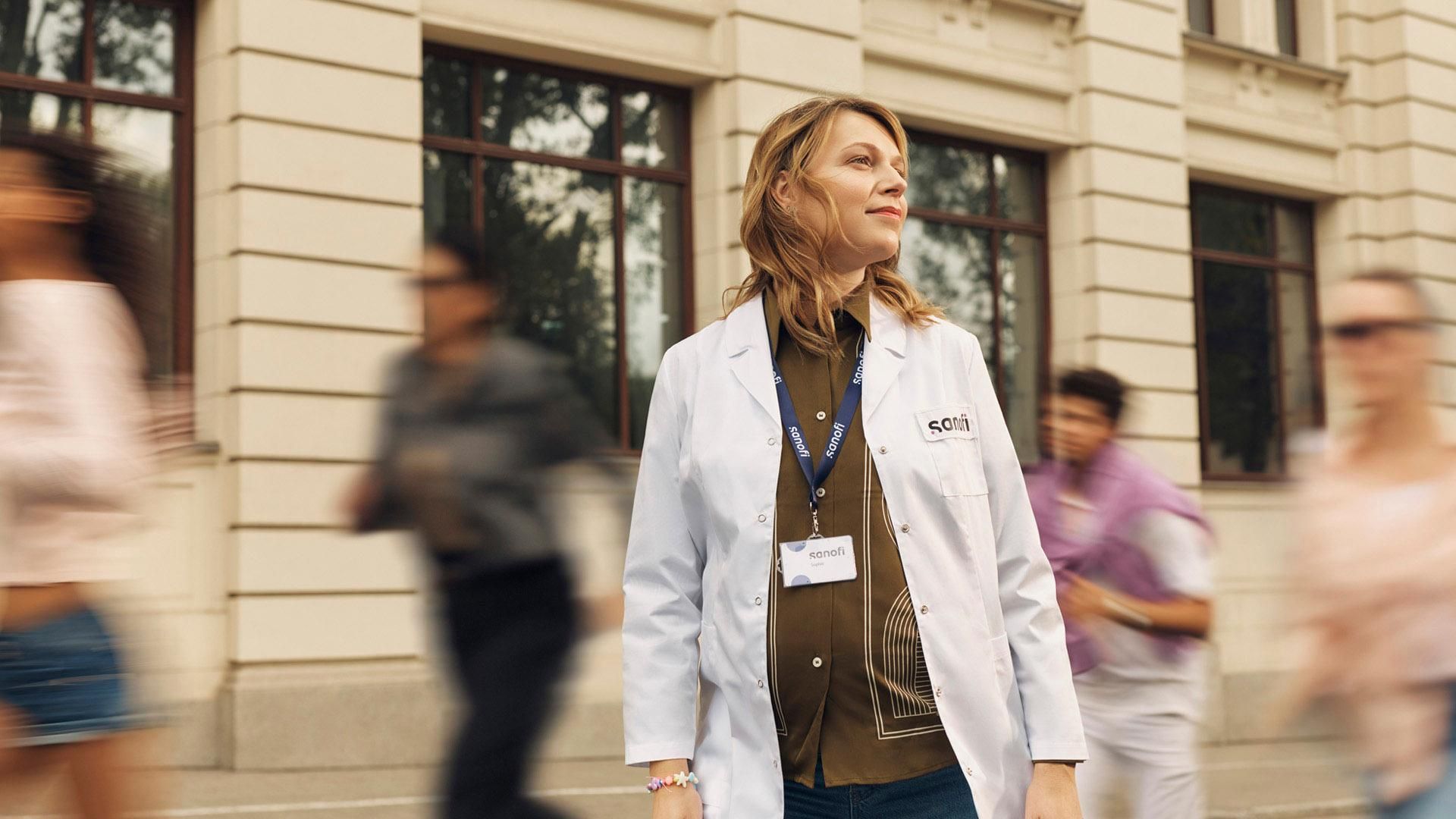A woman in a white Sanofi lab coat and company badge looks ahead with an optimistic expression while standing outside a building. Overlay text reads: "Working in Manufacturing and Supply."​
