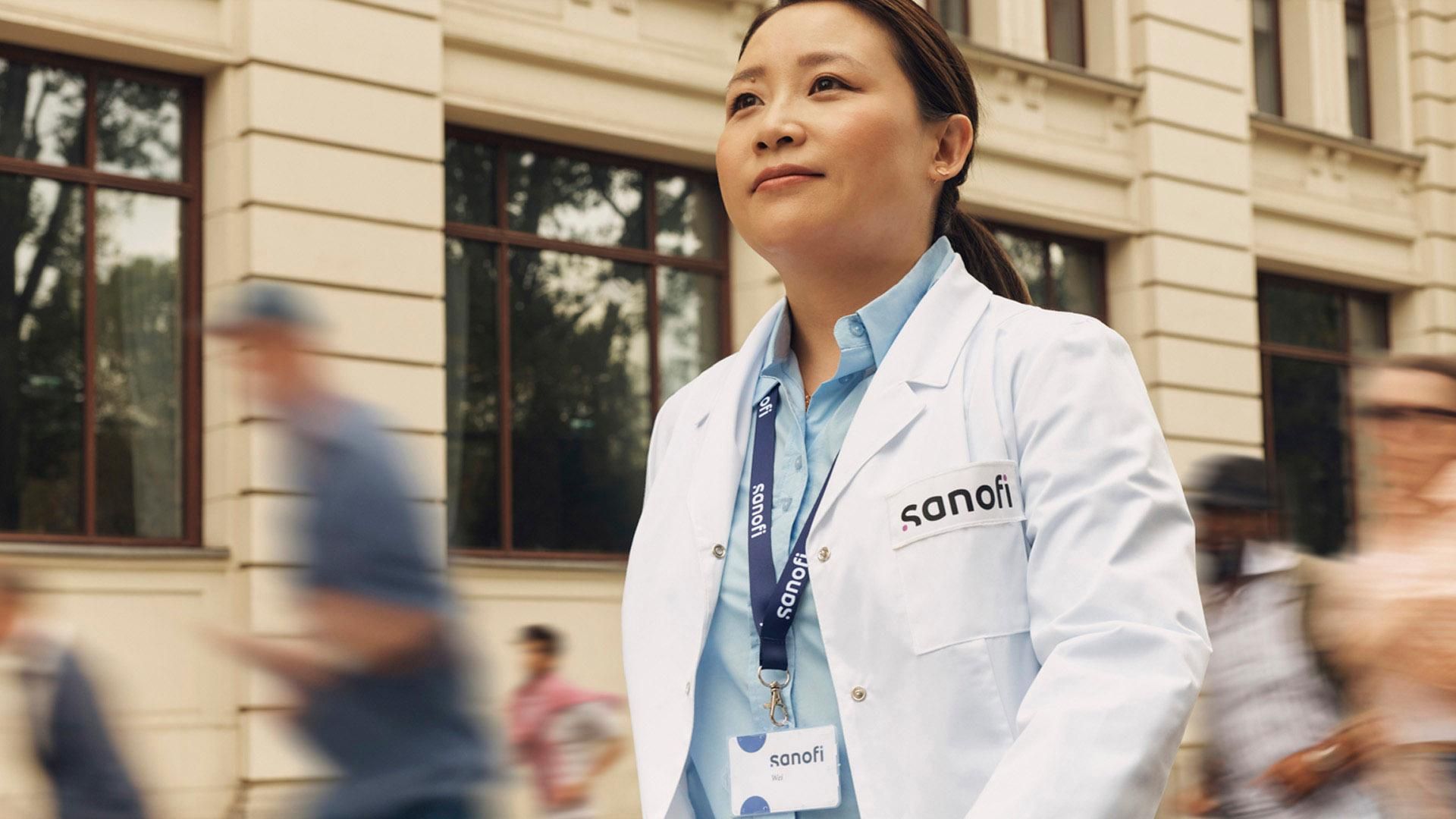 A woman in a white Sanofi lab coat and company badge outside of a building, moving forward while confidently and positively looking ahead. Several people are running alongside her in the background, they are blurred in motion.. Overlay text reads: "Working in Artificial Intelligence."​